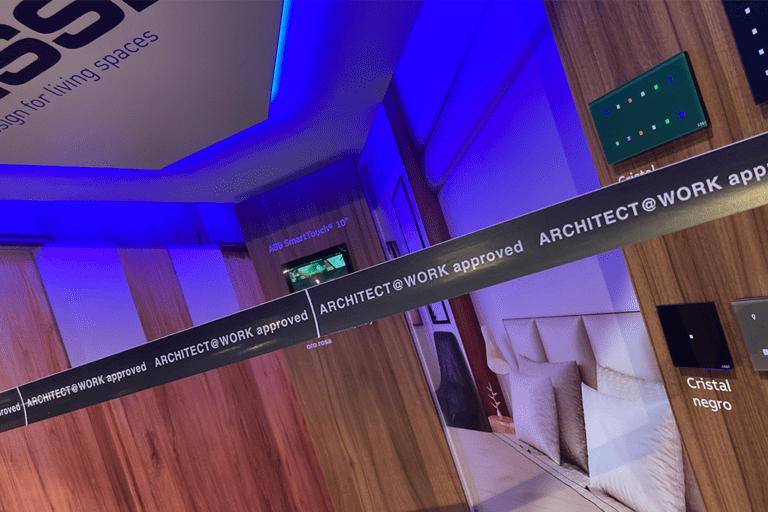 Una vista en ángulo de un stand de exposición con paredes de madera y luces LED azules. La imagen muestra muestras de productos, texto repetitivo que dice "ARCHITECT@WORK approved" y un letrero para un dispositivo "ABB SmartTouch 10"". Se ve una exhibición de dormitorio al fondo.