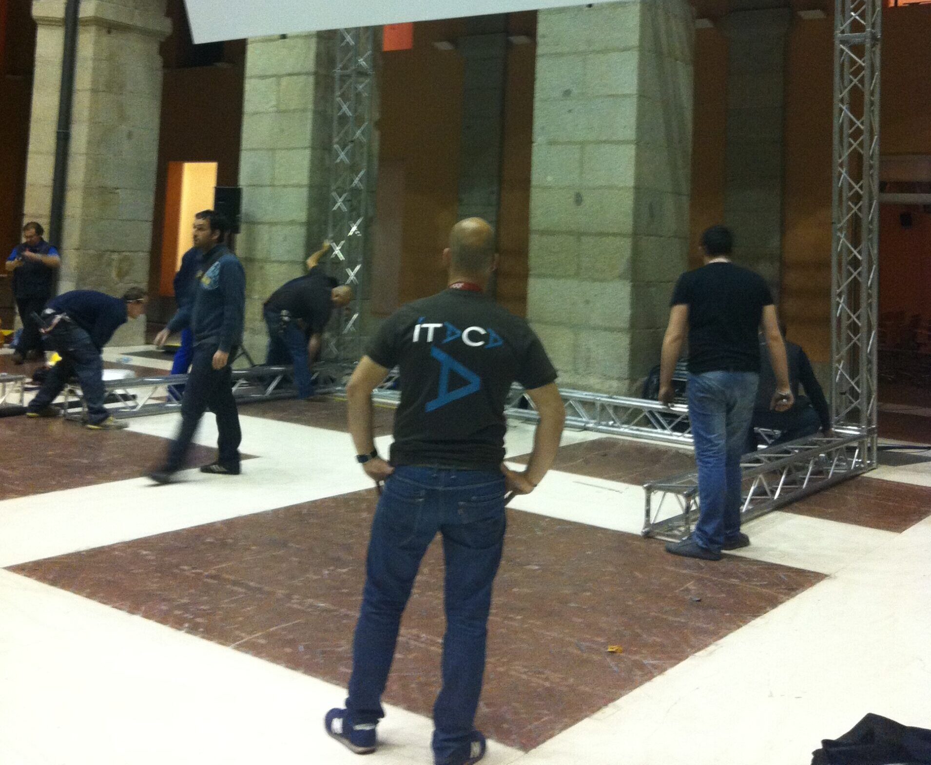 Miembros del equipo de montaje trabajando en la construcción de una gran pantalla y una estructura de truss para un evento, en un espacio interior con arquitectura clásica.