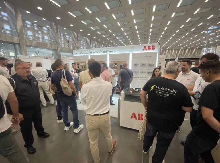 Un concurrido stand de exhibición en una feria comercial o evento. En el fondo, un cartel grande de ABB dice: "Electrificando los edificios del futuro". Varias personas están reunidas alrededor del stand, interactuando y mirando productos eléctricos en exhibición.