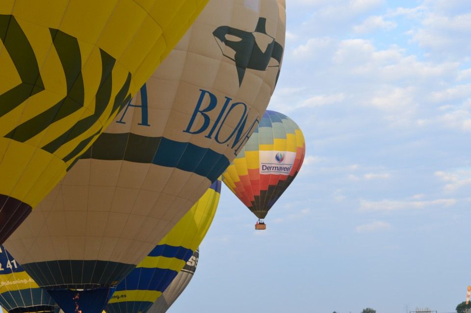 Tres globos aerostáticos se preparan para despegar o están en el aire, vistos desde un ángulo bajo con un cielo azul y algunas nubes como fondo. En primer plano, la parte inferior de un gran globo amarillo con franjas verde oliva y el texto "ABIONE" y "41" visible. Detrás, otro globo de colores brillantes (rojo, naranja, amarillo, azul) lleva el logotipo de "Dermaveel". Los globos parecen ser parte de un evento o festival, con la dirección web "Bedooning.es" también visible cerca de la parte inferior de la imagen.