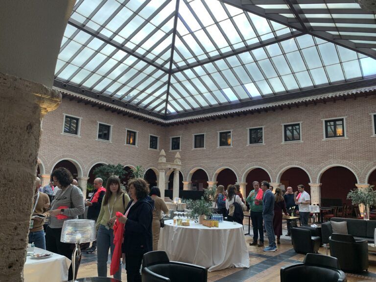 Interior de un antiguo monasterio convertido en el AC Hotel Palacio de Santa Ana en Valladolid, España. La imagen muestra un evento social en el impresionante patio o claustro cubierto, que sirve como área común y restaurante. El espacio conserva la arquitectura original con arcos de ladrillo visto y columnas de piedra, remodelado con un techo acristalado moderno que permite la entrada de luz natural. Varias personas están de pie y charlando alrededor de mesas redondas con manteles blancos, disfrutando del ambiente tranquilo y elegante del hotel de lujo.