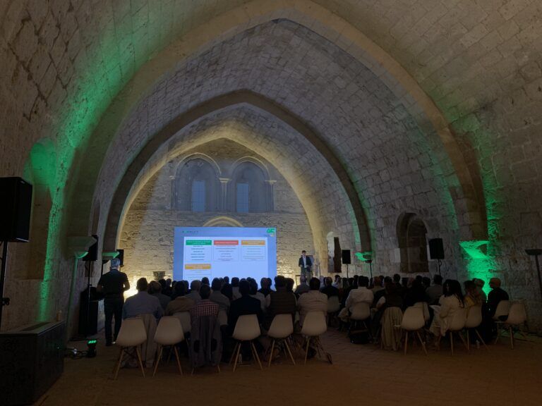 Una presentación durante una conferencia se lleva a cabo en el interior de una sala abovedada de piedra histórica, iluminada con luces verdes. Un orador se encuentra en el escenario frente a una pantalla de proyector, dirigiéndose a una audiencia sentada en sillas blancas. El espacio combina elementos arquitectónicos antiguos con equipamiento moderno para eventos.