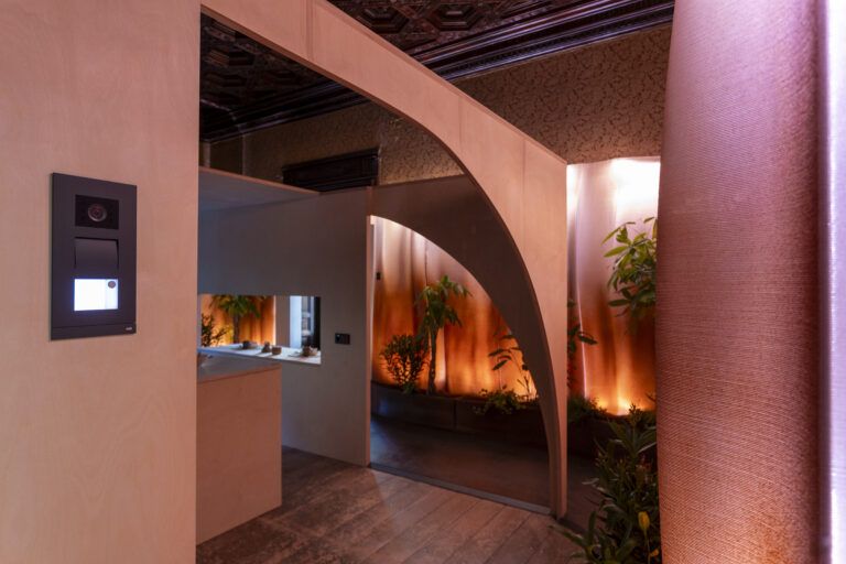 Interior contemporáneo con paneles de madera clara y un sistema de videoportero empotrado en la pared. El espacio combina luz cálida, vegetación interior y formas arquitectónicas curvas.