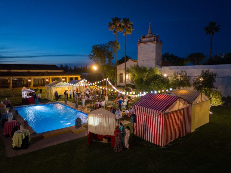 Vista aérea nocturna de una fiesta de estilo andaluz celebrada al aire libre en una finca o hacienda, con una piscina iluminada y varias casetas de feria a rayas rojas y blancas y amarillas. Los invitados socializan bajo hileras de luces de colores, con palmeras y un edificio tradicional español con una torre al fondo.