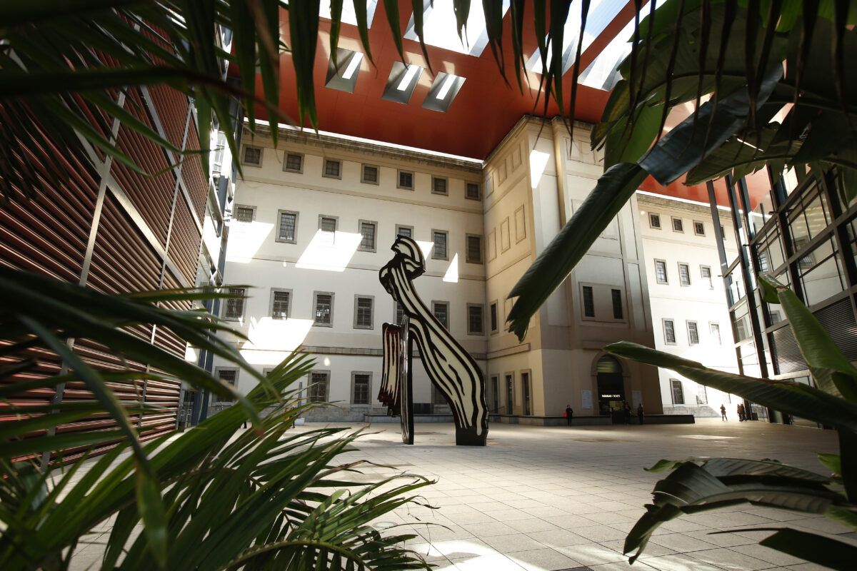 Un patio interior soleado de un edificio con ventanas, enmarcado por grandes plantas verdes en primer plano, contiene una escultura abstracta grande, blanca y negra.