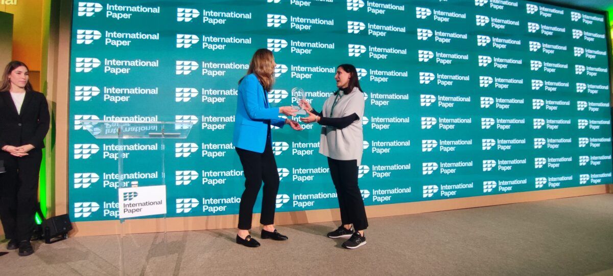 Dos mujeres de negocios de pie en un escenario se dan la mano e intercambian un premio de cristal transparente. La mujer de la izquierda lleva un blazer azul brillante y la de la derecha una blusa blanca y pantalones negros. Detrás de ellas hay una pared verde de fondo cubierta con logotipos repetidos de International Paper.