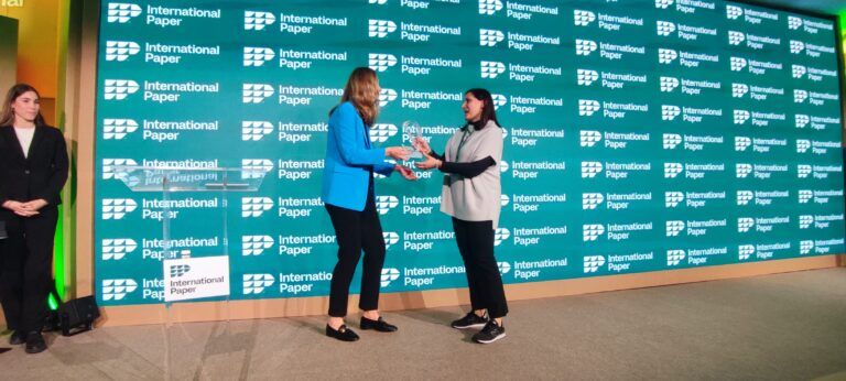 Dos mujeres de negocios de pie en un escenario se dan la mano e intercambian un premio de cristal transparente. La mujer de la izquierda lleva un blazer azul brillante y la de la derecha una blusa blanca y pantalones negros. Detrás de ellas hay una pared verde de fondo cubierta con logotipos repetidos de International Paper.