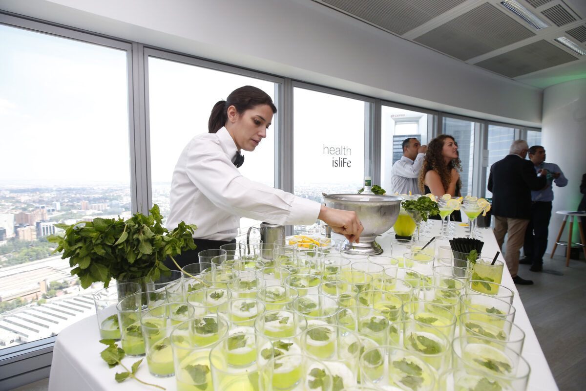 Una camarera preparando cócteles de menta y lima (mojitos) en una barra durante un evento en un rascacielos, con muchas bebidas ya servidas en primer plano y vistas panorámicas de la ciudad a través de las ventanas, donde se lee el texto 'health isliFe.