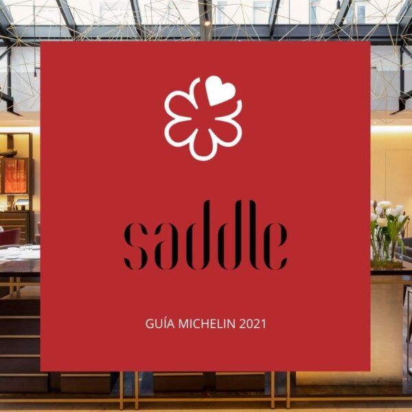 Imagen con fondo rojo que muestra una estrella Michelin blanca y el texto “Saddle – Guía Michelin 2021”. Al fondo se aprecia parte del interior elegante del restaurante Saddle.