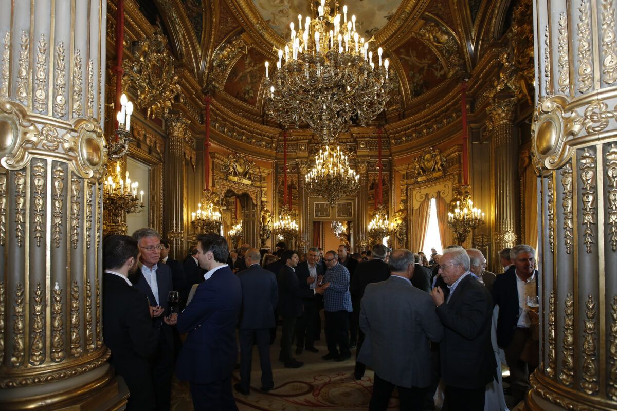 Un salón opulento y ricamente decorado con paredes doradas y grandes lámparas de araña acoge un evento con personas vestidas formalmente. Casa América.