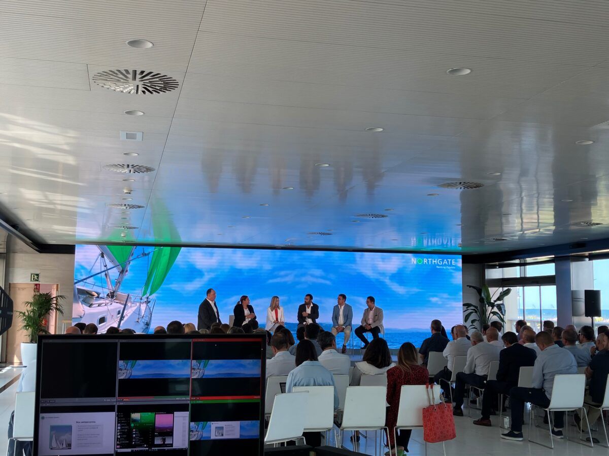 Un evento de conferencia o panel de discusión interior con seis oradores sentados en un escenario frente a una gran pantalla LED que muestra un velero y el logotipo de "NORTHGATE". Una audiencia está sentada en sillas blancas mirando a los oradores. En la esquina inferior izquierda, una estación de control de producción de vídeo con múltiples monitores está en funcionamiento.