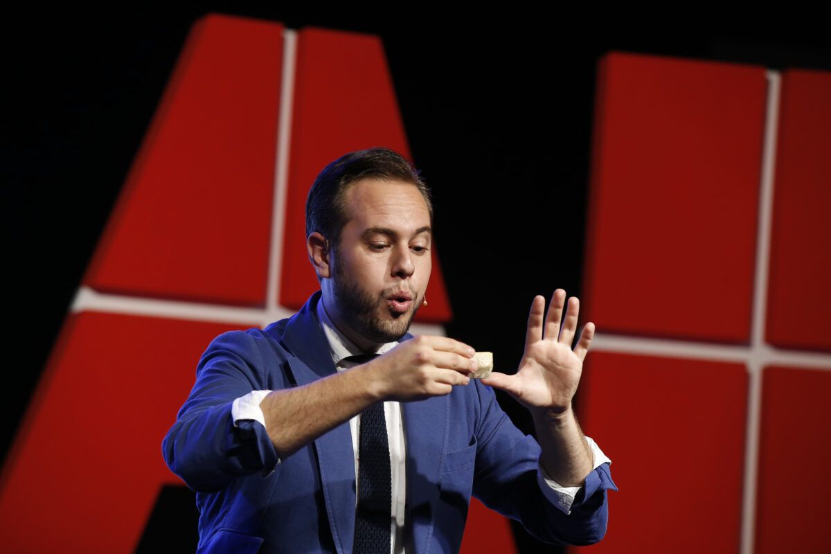 Un hombre con traje azul y corbata en un escenario, sosteniendo un objeto pequeño y gesticulando hace un truco de magia, con un fondo de letras rojas