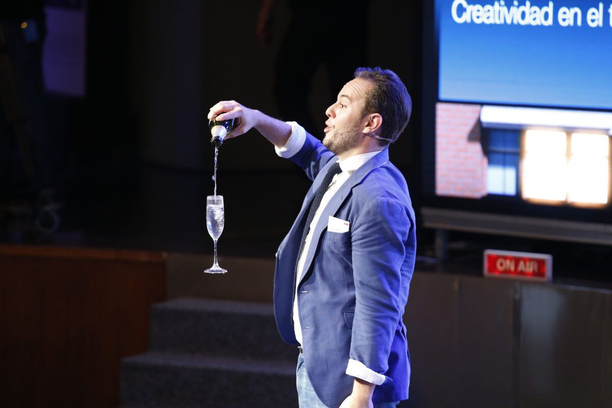 Un hombre vestido con una chaqueta azul vierte champán en una copa de cristal en un escenario, hace un truco de magia.