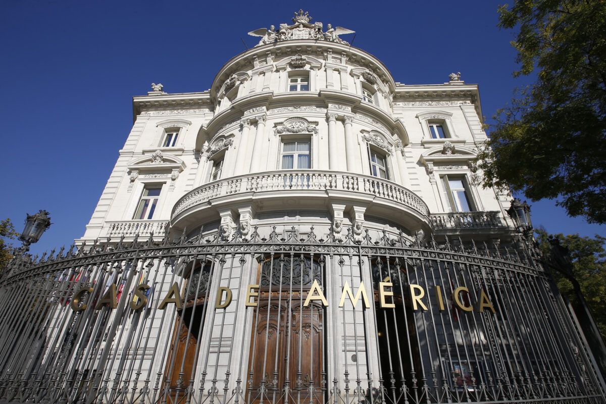 Fachada del histórico Palacio de Linares, sede de la Casa de América, en Madrid. El edificio es de estilo ecléctico, de color blanco, con una sección central curva, balcones con balaustradas y una ornamentación detallada en el tejado. En primer plano, una verja de hierro forjado negra muestra letras doradas que forman la inscripción "CASA DE AMERICA".
