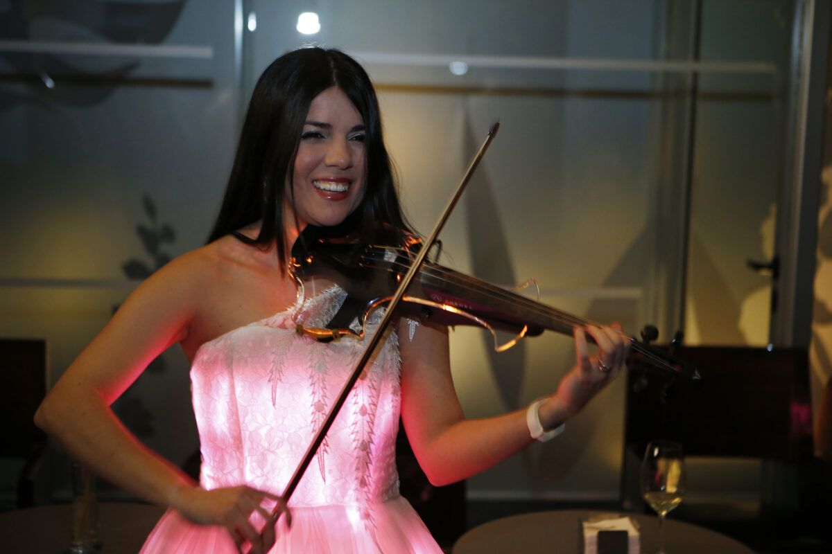 Una mujer sonriente con el pelo negro largo tocando un violín eléctrico iluminado, vistiendo un vestido strapless que brilla con luz rosa.