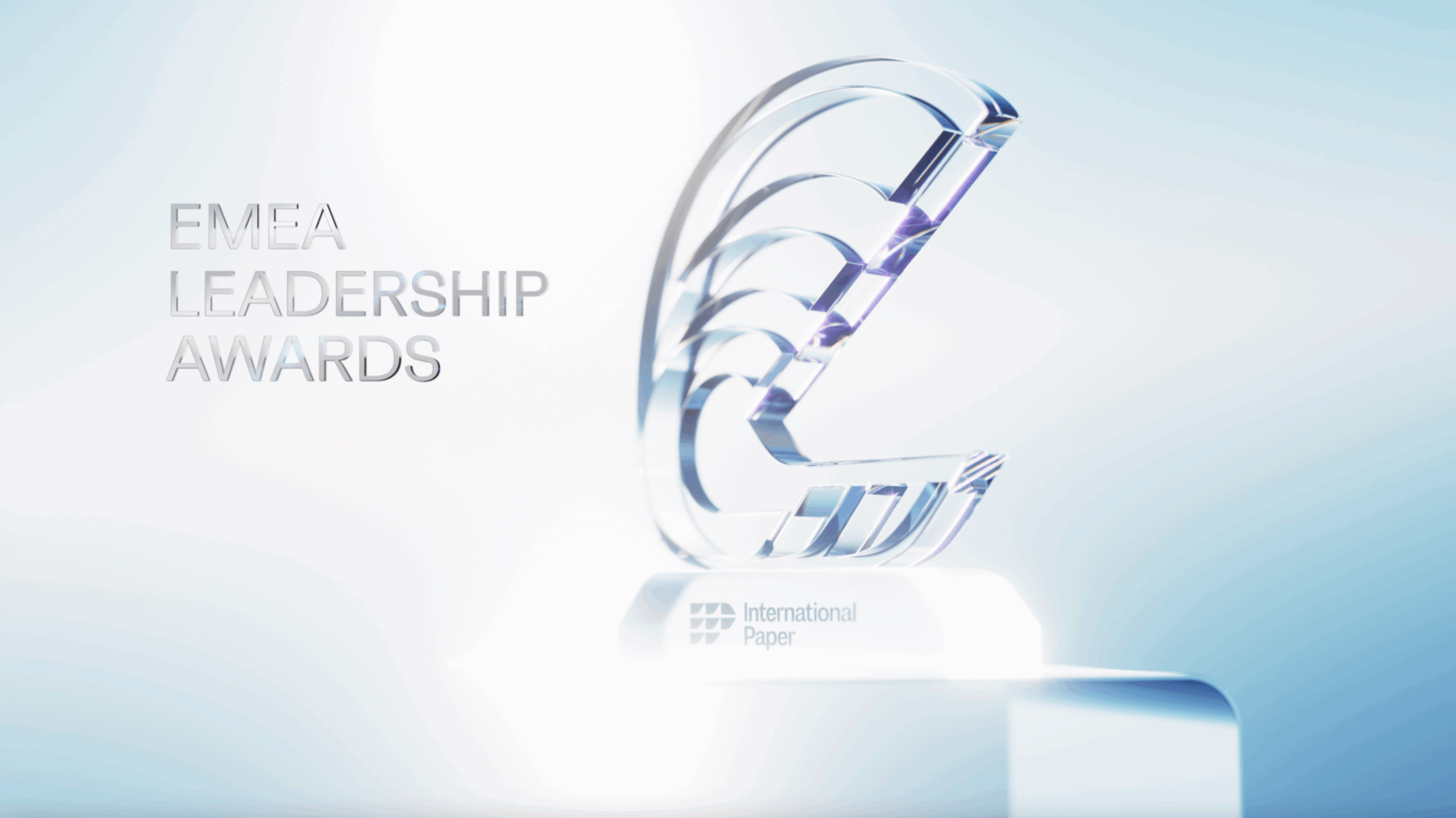 Un trofeo de cristal transparente con un diseño abstracto en forma de C, situado sobre una base iluminada. El texto superpuesto en la imagen dice "EMEA LEADERSHIP AWARDS" y el logotipo y nombre de la empresa "International Paper" están grabados en la base.