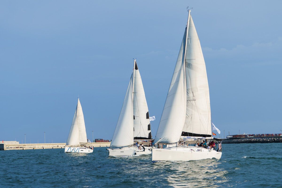 Tres veleros navegan en formación en mar abierto cerca de un rompeolas de cemento, bajo un cielo azul claro. Parece ser una regata o evento de vela, con varias personas visibles en la cubierta de los barcos. La proa del velero en primer plano a la derecha es un Beneteau Oceanis 37, un velero de crucero conocido por su rendimiento y comodidad.