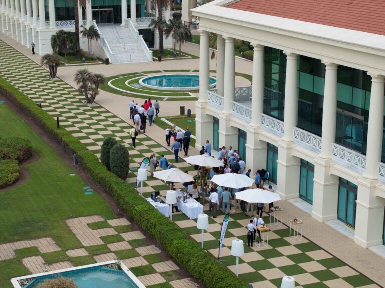 Vista aérea de un evento social o corporativo al aire libre en un resort de lujo de estilo mediterráneo. El complejo cuenta con edificios blancos con columnas, césped verde exuberante, un camino con patrón de tablero de ajedrez, piscinas y palmeras. Numerosas personas están reunidas en la terraza pavimentada, socializando alrededor de mesas y sombrillas blancas.