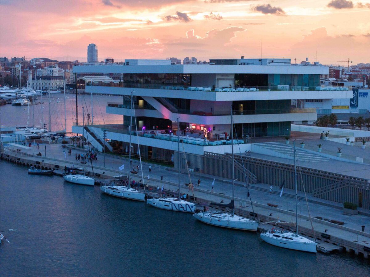 Vista exterior nocturna del Edificio Veles e Vents en la Marina de Valencia, España, al atardecer, con un cielo rosado y morado. El edificio moderno de cuatro plantas, diseñado por David Chipperfield, presenta amplias terrazas en voladizo con barandillas de cristal y una iluminación arquitectónica blanca y morada. En la dársena, varios veleros están amarrados a lo largo del muelle en primer plano. Hay actividad en las terrazas, con gente socializando en lo que parecen ser áreas de restaurante o eventos. La imagen tiene una marca de agua de texto que dice "Seralla".