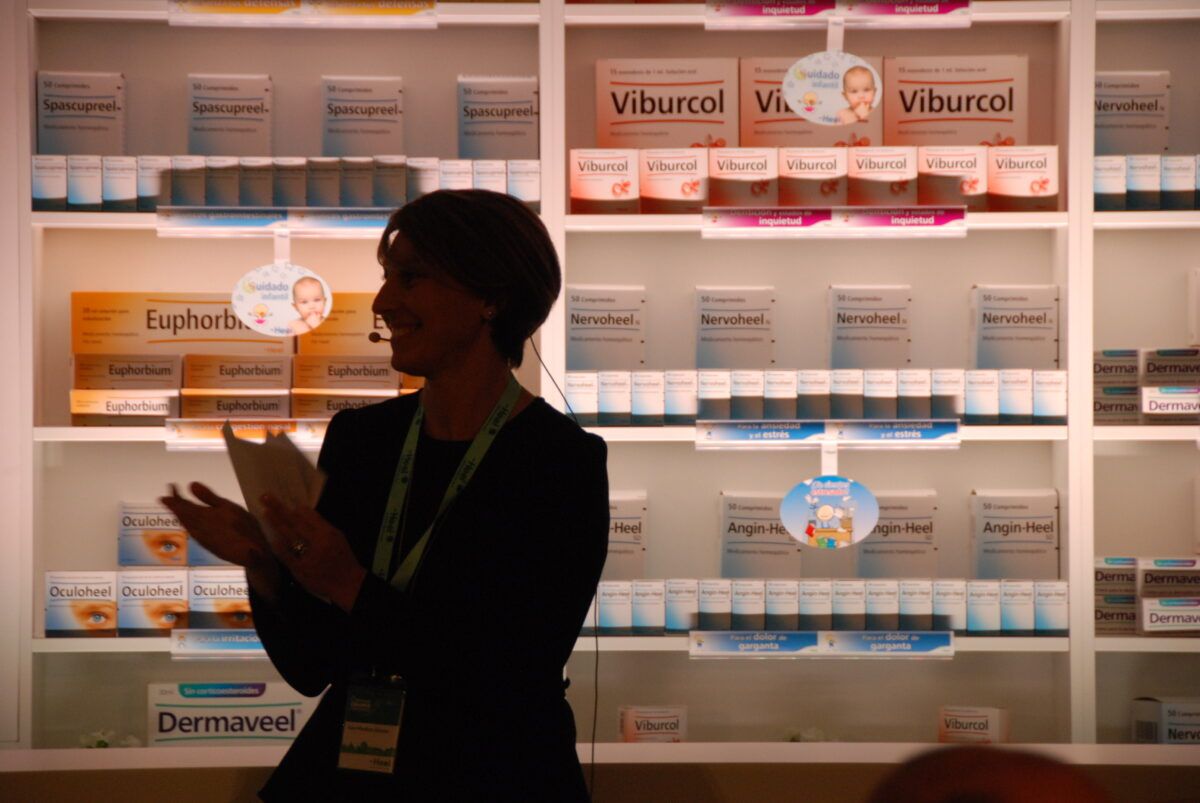 Una mujer de perfil, vestida de oscuro, hablando y gesticulando en un evento corporativo o feria comercial. Está de pie frente a un gran expositor de pared blanco lleno de productos farmacéuticos de la marca Heel, incluyendo cajas con nombres como Spascupreel, Viburcol, Nervoheel, Euphorbium y Dermaveel
