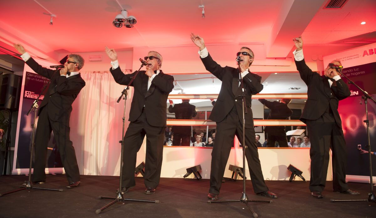 Cuatro hombres vestidos con trajes negros y gafas de sol actúan en un escenario con iluminación roja en un evento corporativo, con el logotipo de Niessen visible en el fondo.
