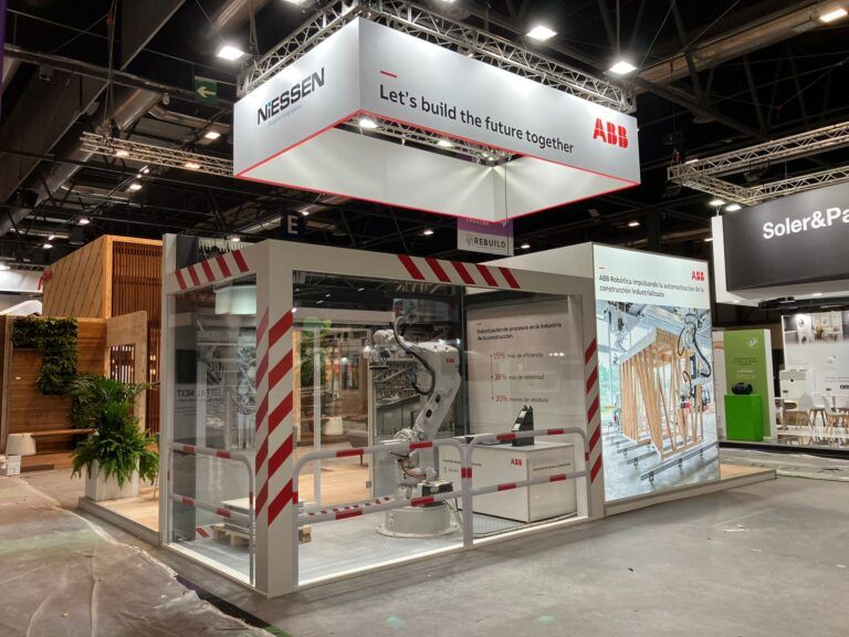 Un stand de exposición de ABB y Niessen en un evento, mostrando un robot industrial blanco dentro de una vitrina de cristal, con el lema "Let's build the future together" en un banner colgante.