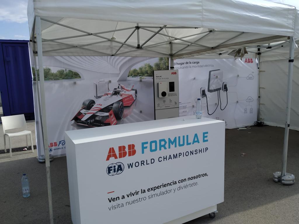Un stand promocional para el Campeonato Mundial ABB FIA Fórmula E en un evento al aire libre. El stand blanco cuenta con un gran fondo que muestra un coche de carreras eléctrico Mahindra Racing rojo y blanco y cargadores de vehículos eléctricos ABB Terra AC. En primer plano hay un mostrador con el logotipo de ABB Fórmula E y un eslogan que invita a los visitantes a probar un simulador.