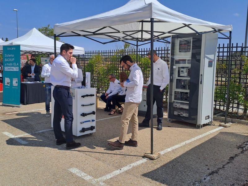 Un evento o feria comercial al aire libre donde profesionales de la industria solar se reúnen bajo carpas blancas. La escena se centra en un stand de la marca española Loscano, que exhibe un sistema compacto de control de respaldo (backup box) para instalaciones fotovoltaicas y un armario eléctrico más grande. Un cartel azul con el texto "Loscano Sistema BACKUP y modo ISLA COMPACTO" es visible a la izquierda. Varias personas vestidas de forma profesional están interactuando y observando los productos relacionados con la energía solar y los sistemas de respaldo.