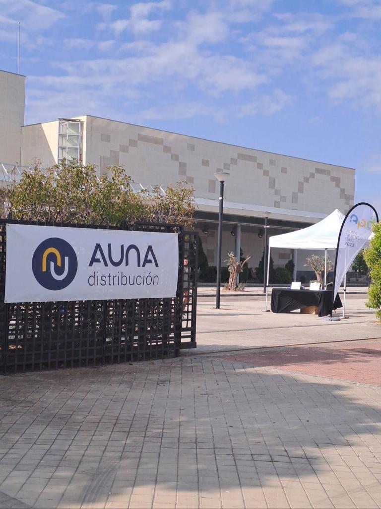 Un cartel grande para AUNA distribución cuelga de una valla negra en el exterior, con el logotipo de la compañía. En el fondo, se ve un edificio moderno de color beige bajo un cielo parcialmente nublado. A la derecha, una carpa blanca para eventos con una mesa negra debajo y una bandera de vela más pequeña con el texto "AUNA PARTNER DAYS 2023" es visible.