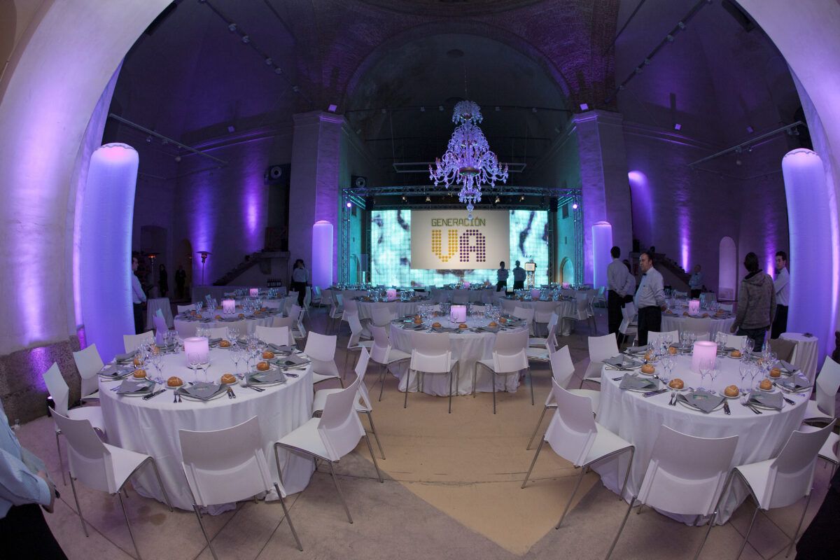 Un gran salón abovedado está preparado para un evento formal con iluminación ambiental púrpura. Varias mesas redondas vestidas con manteles blancos y sillas blancas ocupan la sala. Varias personas están de pie o sentadas en el lugar.
