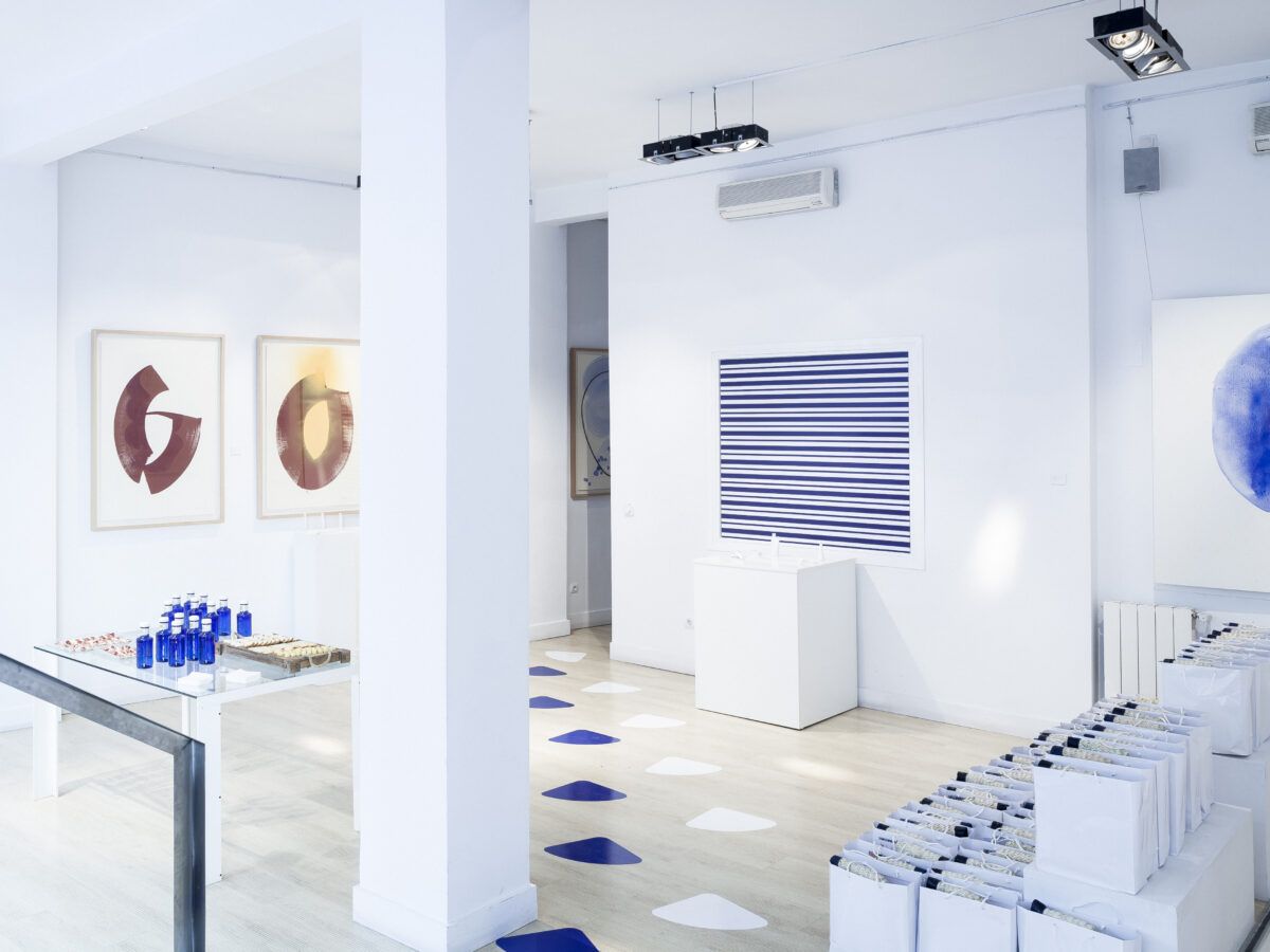 Interior minimalista y luminoso de una galería de arte o espacio de exposición con paredes blancas y suelo de madera clara. Se muestran varias obras de arte abstracto, incluyendo dos cuadros con formas marrones a la izquierda, una obra a rayas azules y blancas en el centro y una pintura circular azul a la derecha. La sala está decorada con botellas azules, bolsas blancas y huellas azules y blancas que marcan un camino en el suelo.