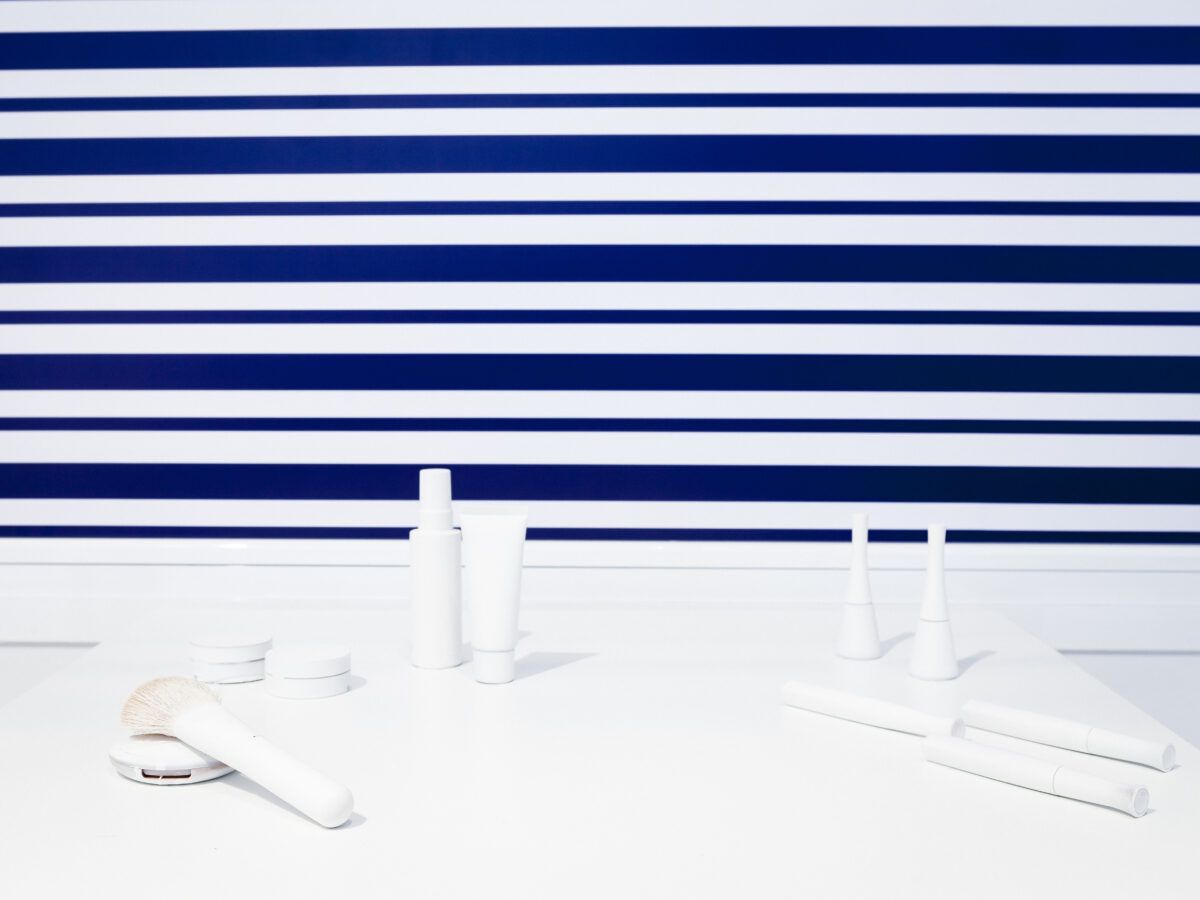 Arreglo de productos cosméticos blancos sin marca, que incluyen envases, un tubo, dos pequeños botes cónicos y una brocha de maquillaje, sobre una superficie blanca frente a un fondo de rayas horizontales azules y blancas.