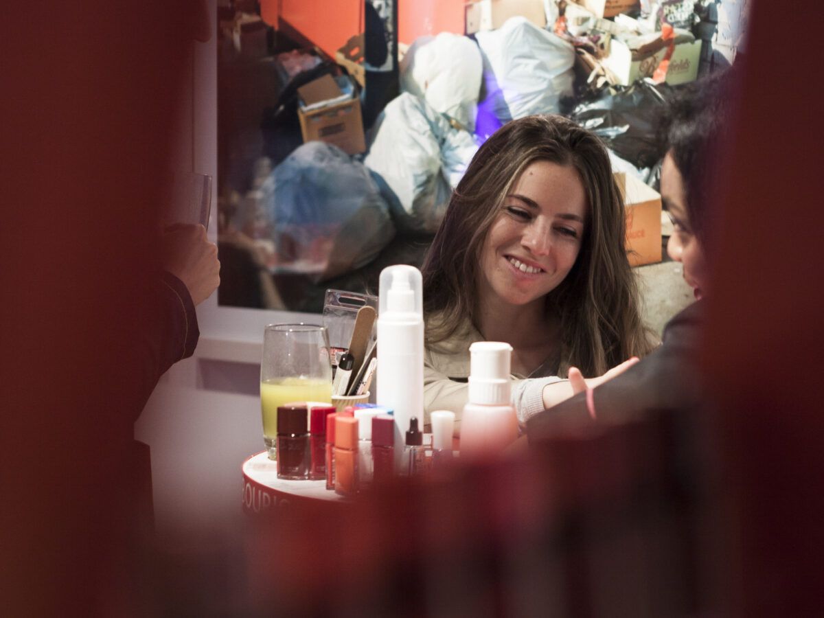 Dos mujeres socializando en un evento, sentadas en una mesa circular con varios productos de belleza o cuidado de la piel. Una de ellas sonríe mientras mira hacia la otra.