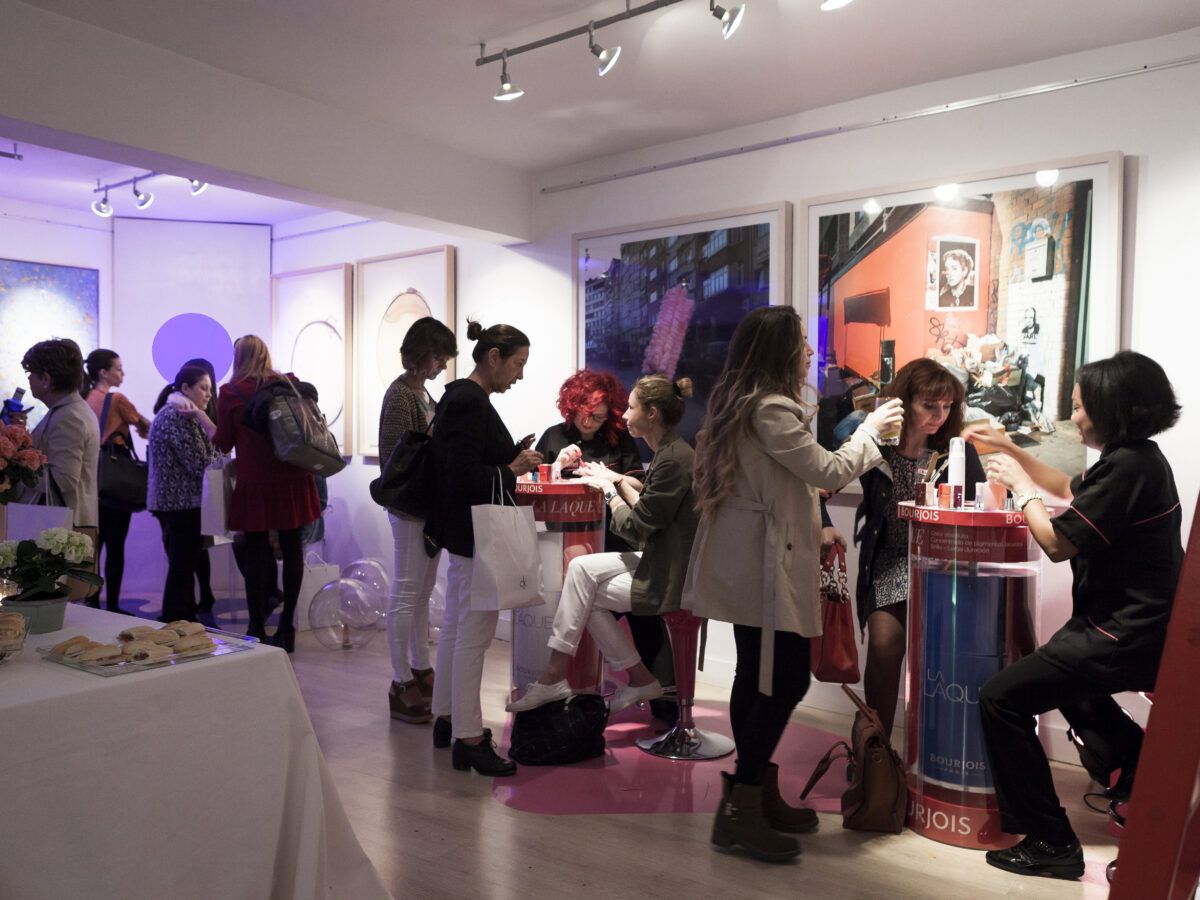 Invitados socializando y comprando productos en el stand de Bourjois en un evento. Varias personas interactúan en torno a una barra circular y una mesa con cosméticos; se observan obras de arte con iluminación morada en las paredes.