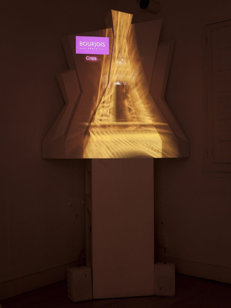 Una instalación o stand de exhibición de la marca Bourjois Paris, con una proyección de luz dorada que simula la Torre Eiffel sobre una estructura geométrica blanca y un cartel morado que dice 'BOURJOIS PARIS' y 'Crea...