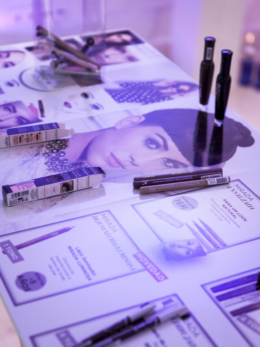 Una mesa de exposición iluminada con luz morada, mostrando productos de maquillaje, incluyendo lápices delineadores, cajas de productos e impresiones de modelos con maquillaje.