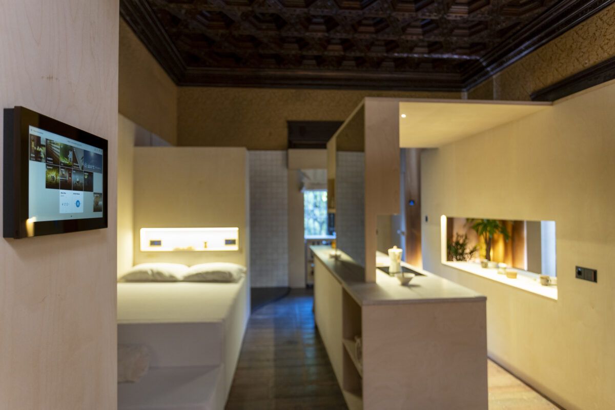 Dormitorio y zona de estar de diseño minimalista, con panel táctil en la pared para controlar funciones domóticas. Los tonos neutros y la iluminación suave crean un ambiente acogedor.