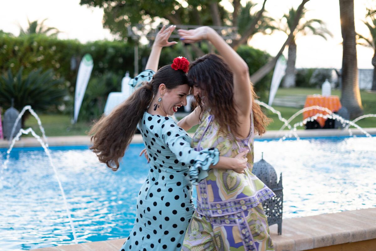 Dos mujeres bailando animadamente al aire libre junto a una piscina con fuentes. Una mujer lleva un vestido de lunares y una flor roja en el pelo; la otra lleva un vestido estampado. El fondo sugiere un evento o fiesta con palmeras y zonas de jardín.