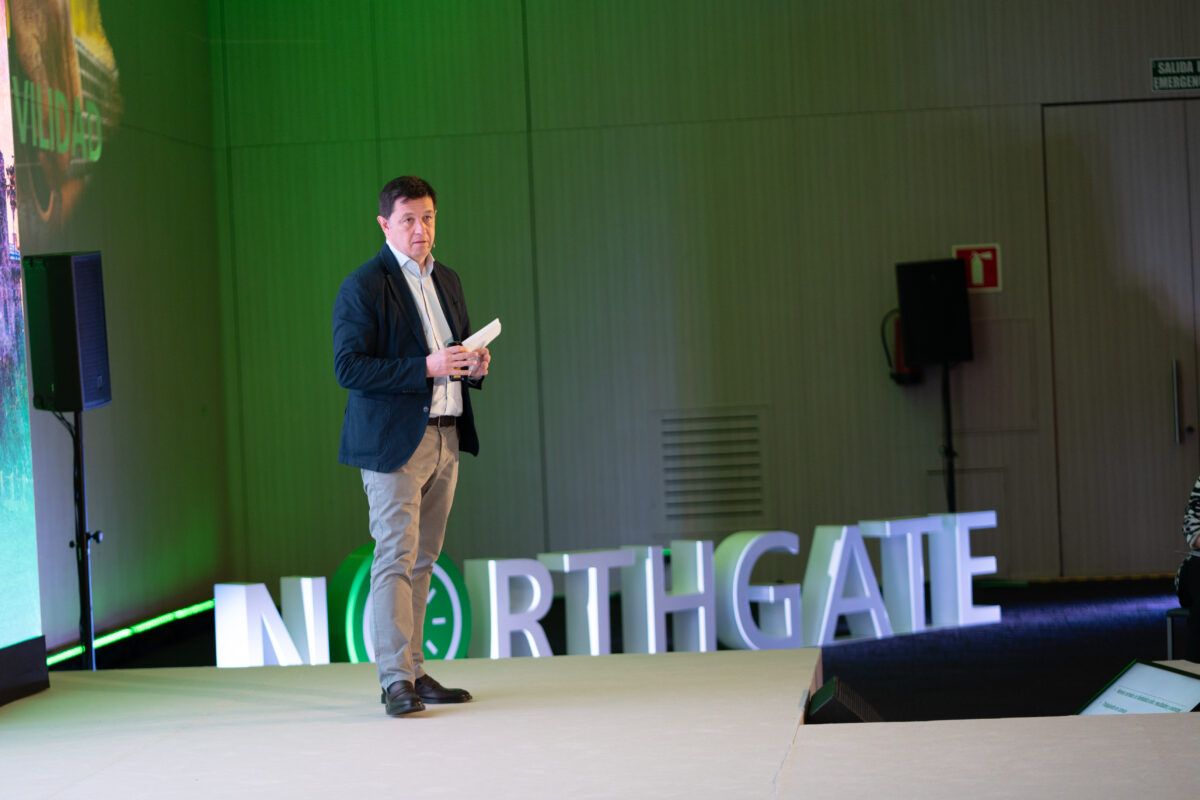 Un orador vestido con chaqueta y pantalón gris de pie en un escenario iluminado, con el logotipo grande de Northgate detrás. El ponente sostiene papeles y un micrófono, y se observa un cartel de 'SALIDA EMERGENCIA' en el fondo, indicando un evento corporativo o conferencia.