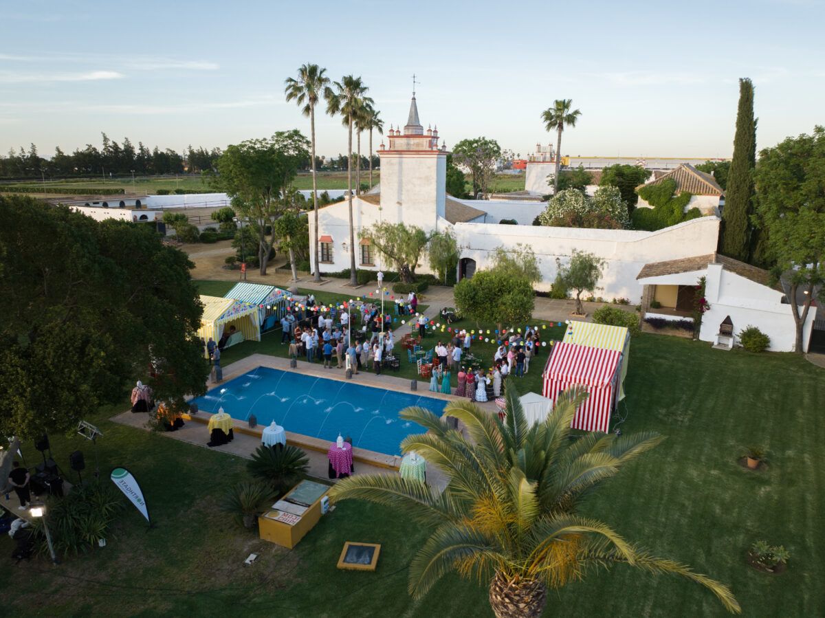Vista aérea de un evento social o fiesta al aire libre en una hacienda o finca con edificios blancos, palmeras y una piscina con fuentes, con invitados socializando entre carpas a rayas amarillas y rojas.