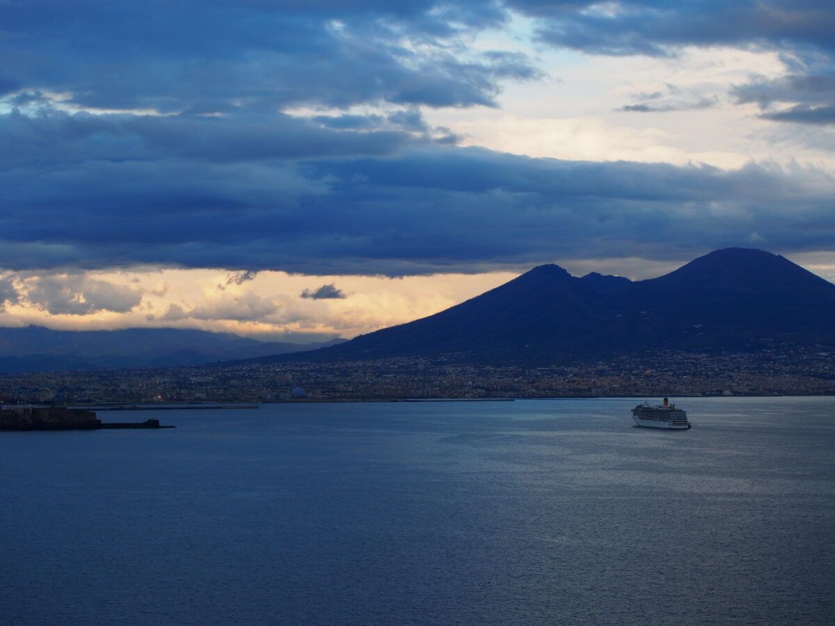 Una vista panorámica de la bahía de Nápoles al atardecer, con el monte Vesubio como telón de fondo, un crucero navegando en primer plano y un cielo dramático y nublado.