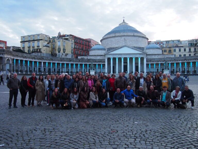 Un grupo grande de personas posa para una foto grupal de noche frente a la Basílica Real de San Francisco de Paula en la Piazza del Plebiscito de Nápoles, cuyas columnas están iluminadas con luces azules.