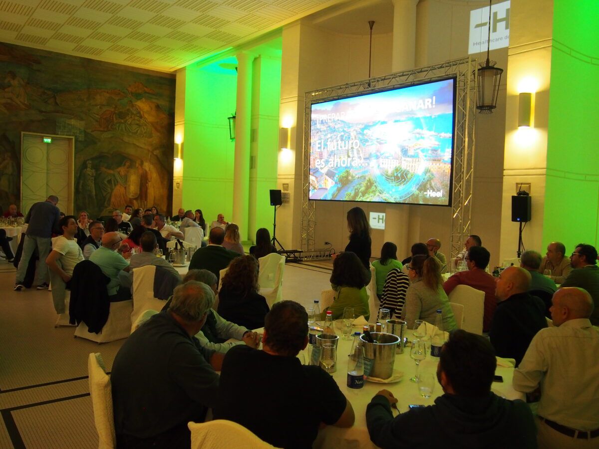 Un evento de conferencia o gala interior bien iluminado en verde, con asistentes sentados en mesas redondas frente a un escenario con una gran pantalla de proyección que muestra el texto: "El futuro es ahora..."