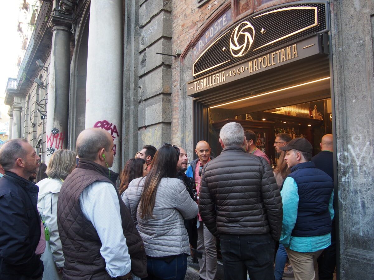 Un grupo de personas se agrupa frente a la entrada de una tienda llamada "R.R. POSTE - TARALLERIA - 1940 - NAPOLETANA" en un edificio de piedra con columnas.