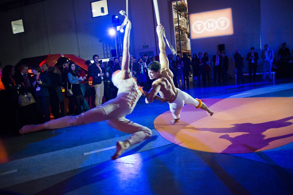 Dos acróbatas, un hombre y una mujer, realizan una rutina con telas aéreas suspendidos en un evento interior iluminado con luces azules y naranjas, con una audiencia de pie en el fondo.