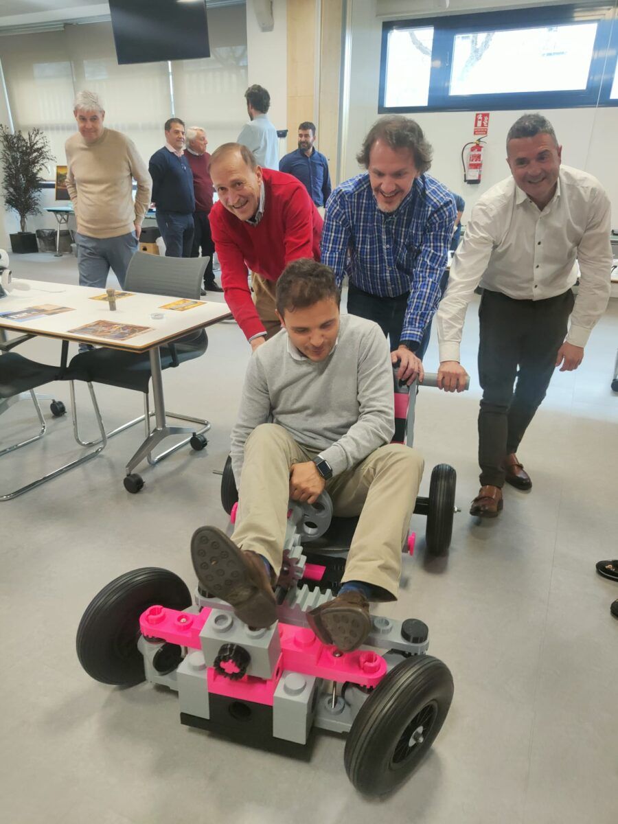 Un hombre sentado en un vehículo de bloques de construcción tipo Lego rosa y gris, siendo empujado por otros tres hombres que sonríen, en lo que parece ser un evento de team building en una oficina