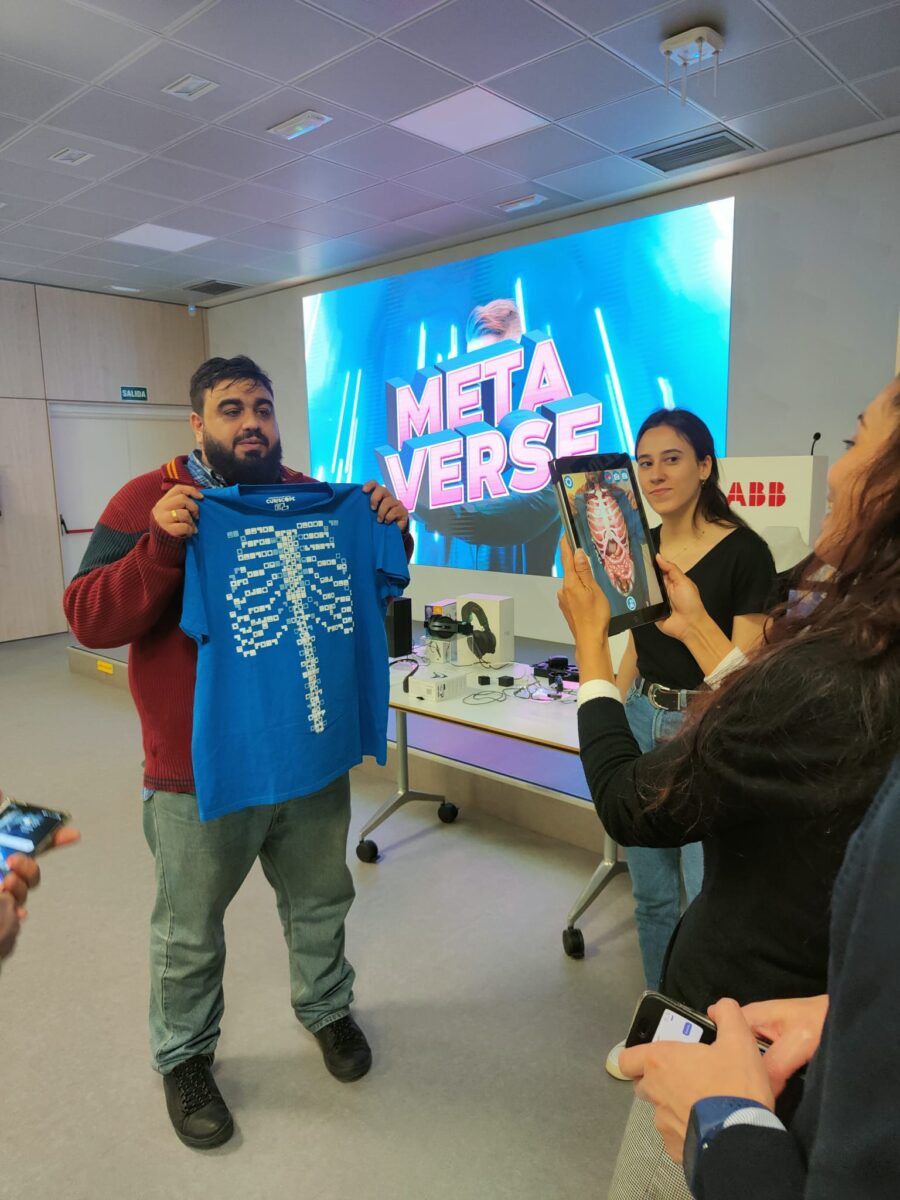 Un hombre sostiene una camiseta azul con un diseño de esqueleto, mientras una persona a su lado apunta una tableta a la camiseta, que muestra una superposición de realidad aumentada del sistema circulatorio humano. La escena tiene lugar frente a una pantalla grande con las palabras 'METAVERSE' y 'ABB' escritas en ella.