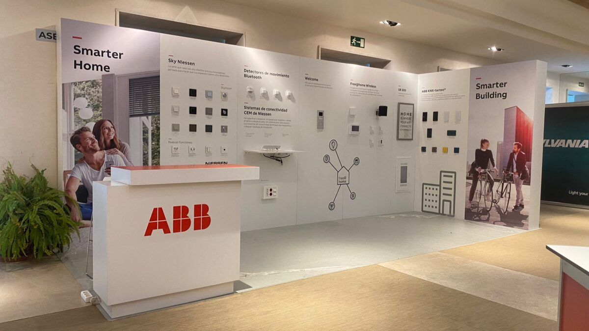 Un stand de exhibición interior para ABB con señalización de "Smarter Home" y "Smarter Building". El stand presenta varios interruptores, detectores de movimiento Bluetooth, sistemas de conectividad de Niessen y otros productos para el hogar inteligente. Incluye un mostrador de recepción con el logotipo de ABB y gráficos de estilo de vida con personas.