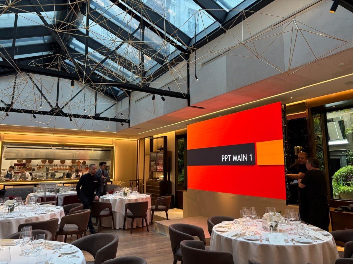 Interior del restaurante Saddle con mesas redondas preparadas para el servicio, una gran pantalla encendida que muestra “PPT MAIN 1”, y varios empleados trabajando cerca de la cocina abierta.