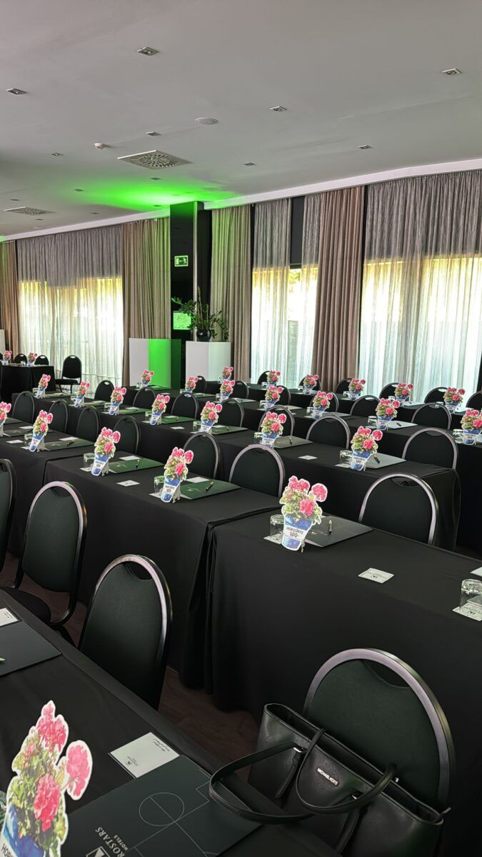Sala de conferencias de Eurostars Hotels montada para un evento, con filas de mesas con manteles negros, sillas oscuras y carpetas decoradas con un cartel de geranios rosas.